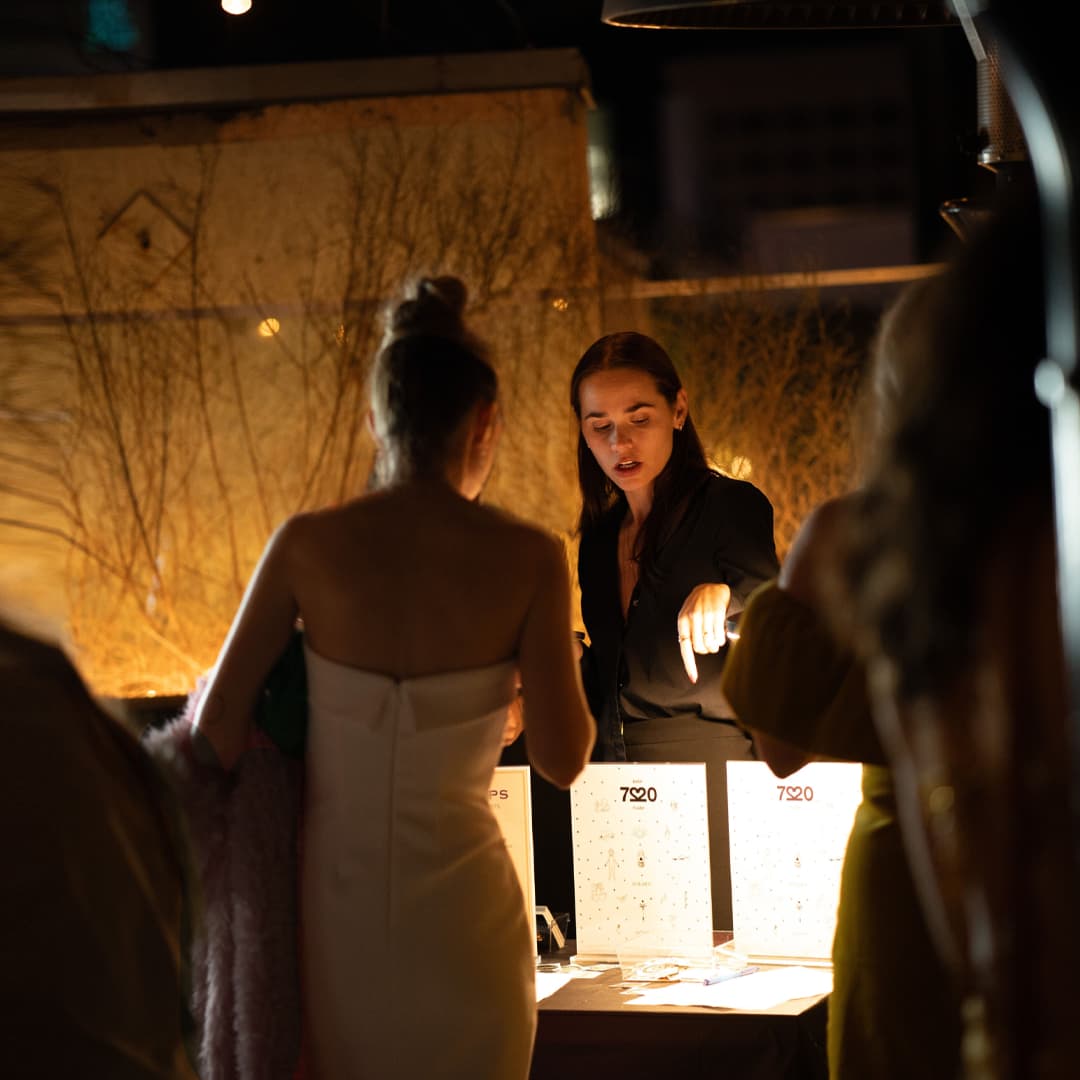 Woman in black talks to a woman in white dress at a dimly lit table with illuminated displays at night.