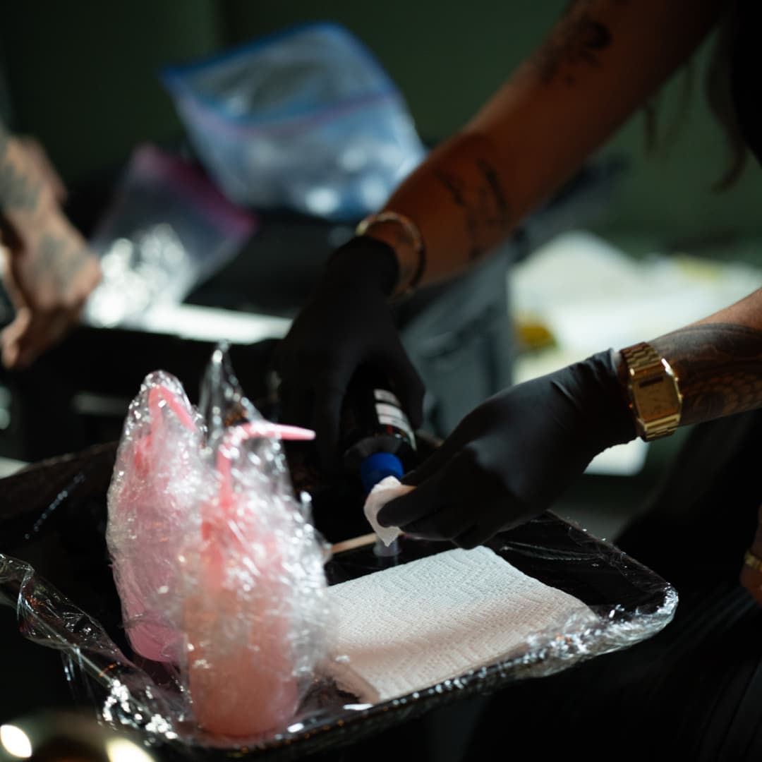 Tattoo artist wearing black gloves preparing ink on a plastic-wrapped tray with supplies nearby.