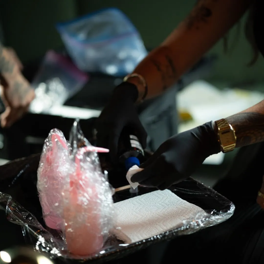 Tattoo artist wearing black gloves preparing ink on a plastic-wrapped tray with supplies nearby.