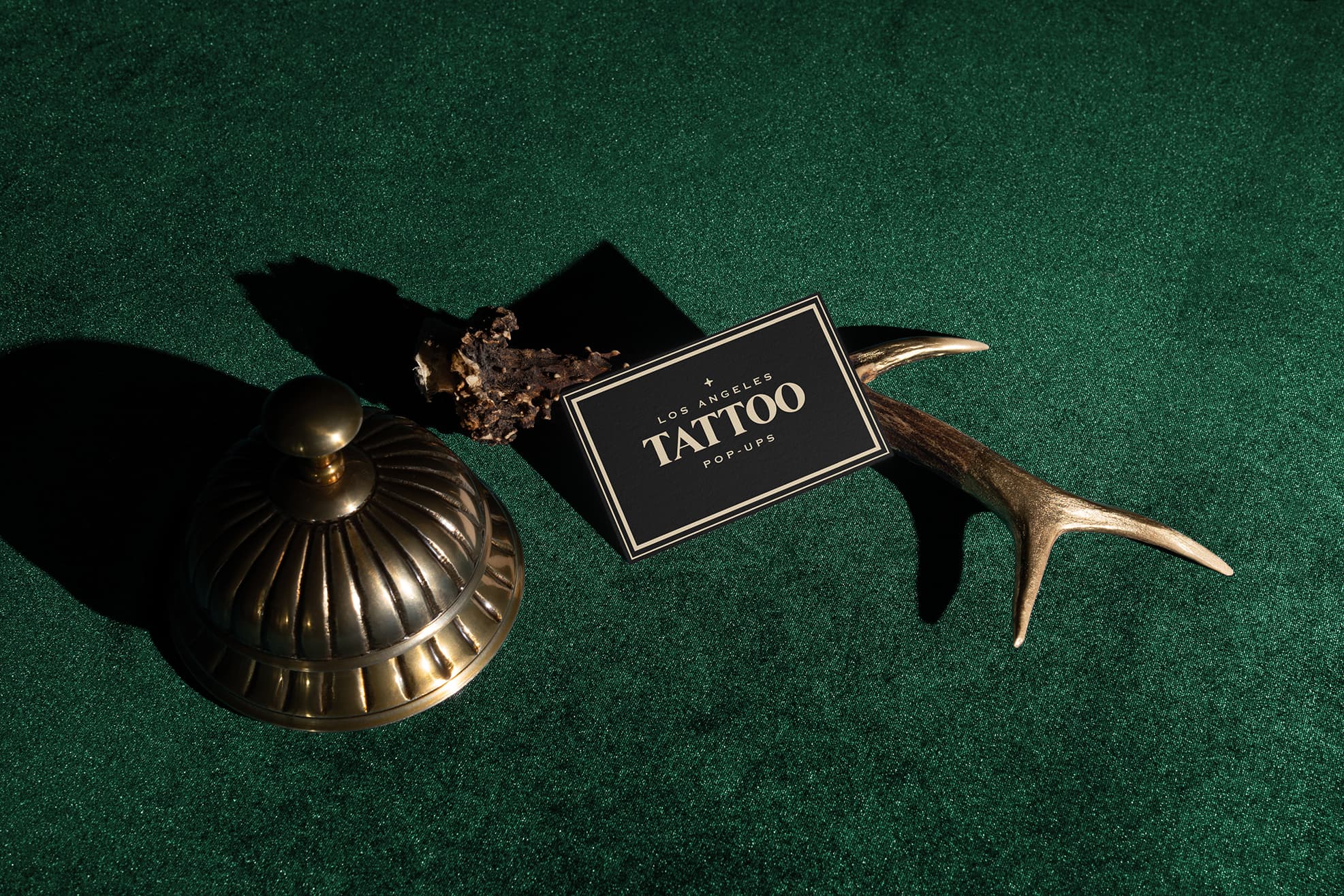Brass bell and antler holding a black card that reads Los Angeles Tattoo Pop-Ups on green surface.