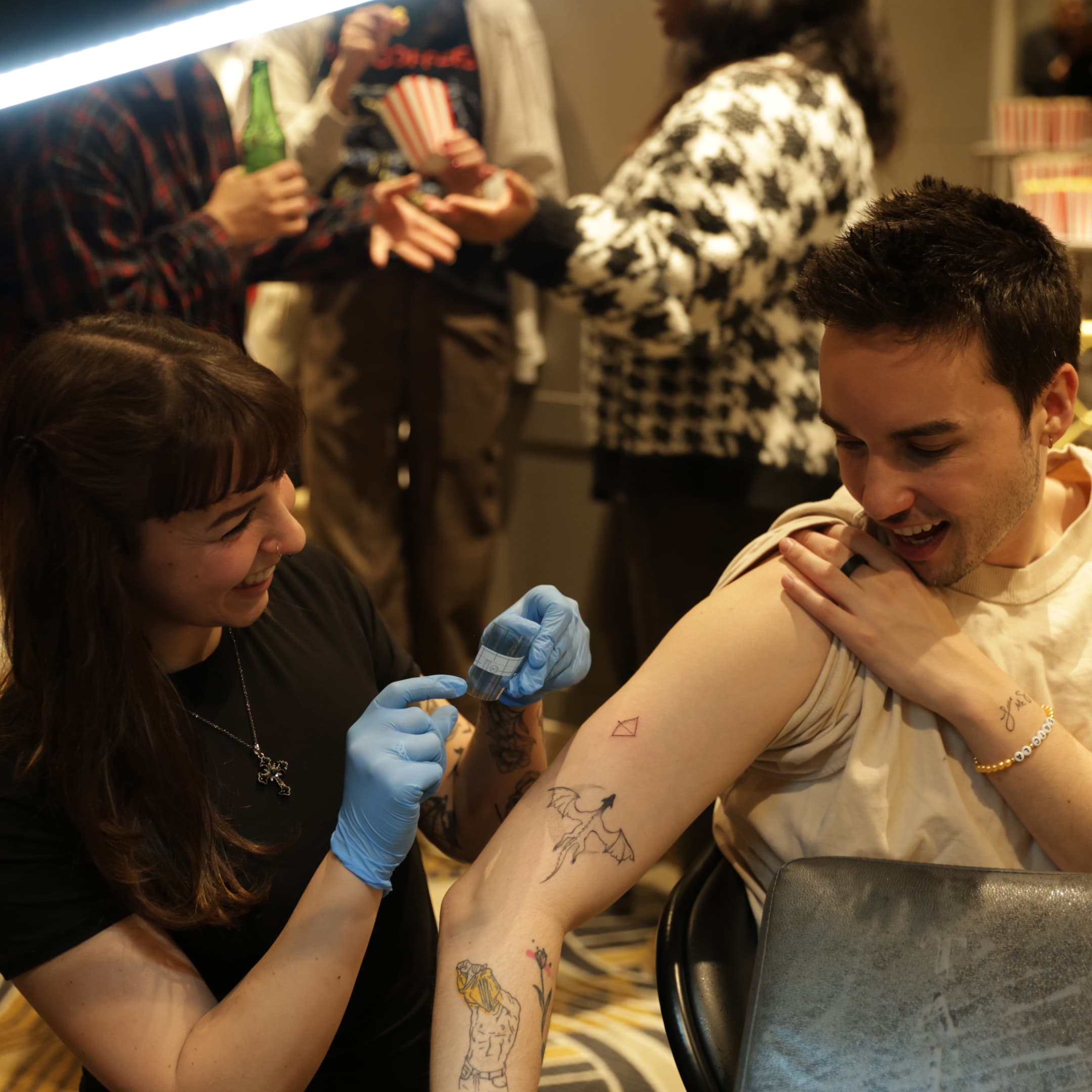 Smiling tattoo artist in blue gloves applies a tattoo on a seated man's upper arm at a social event.
