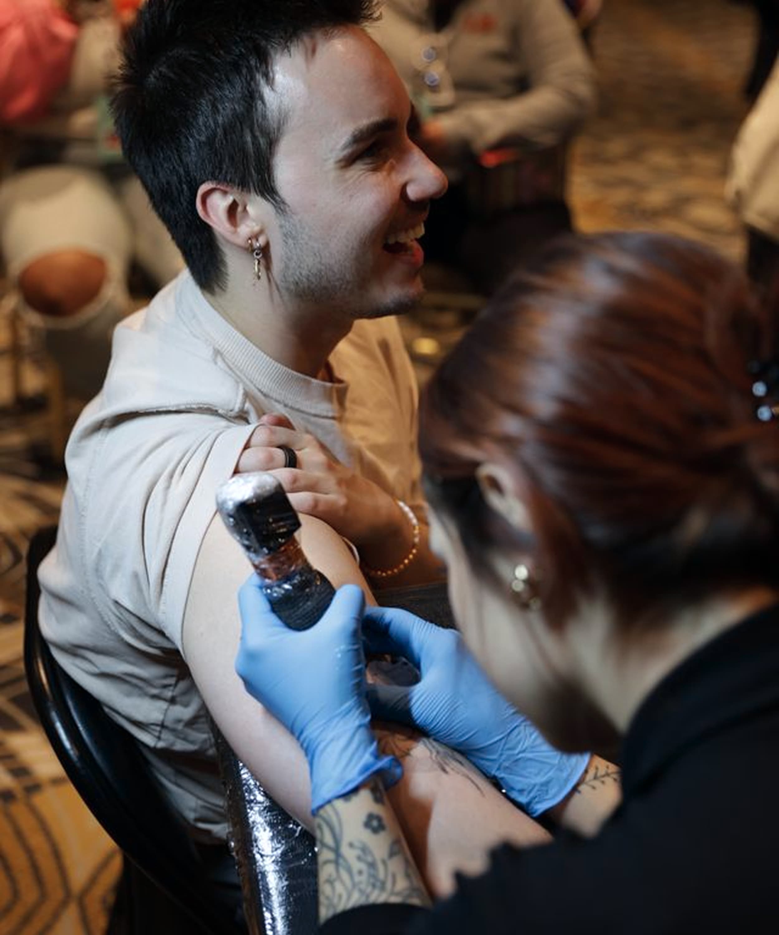 Tattoo artist with blue gloves working on a smiling man's upper arm tattoo.