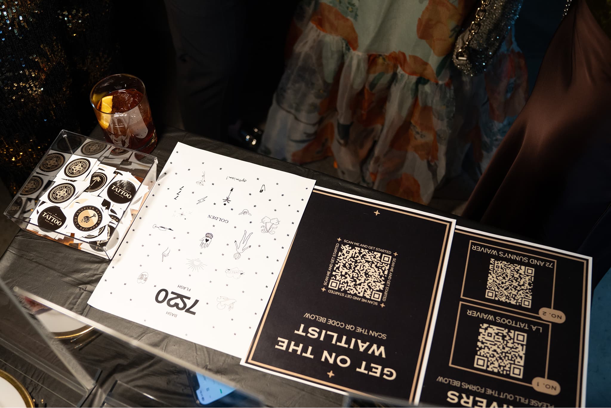 Table with sticker cards, a drink, and signs featuring QR codes for a waitlist and tattoo giveaways.