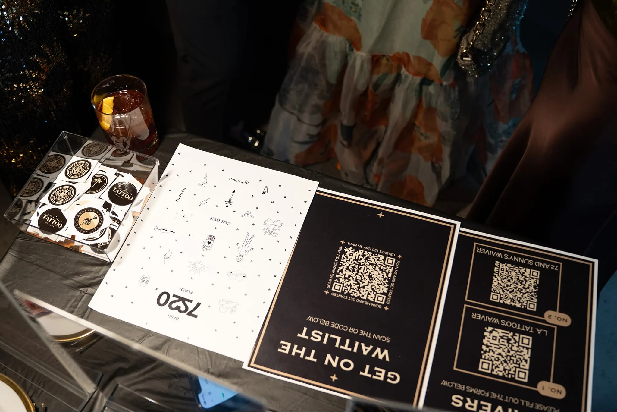 Table with sticker cards, a drink, and signs featuring QR codes for a waitlist and tattoo giveaways.