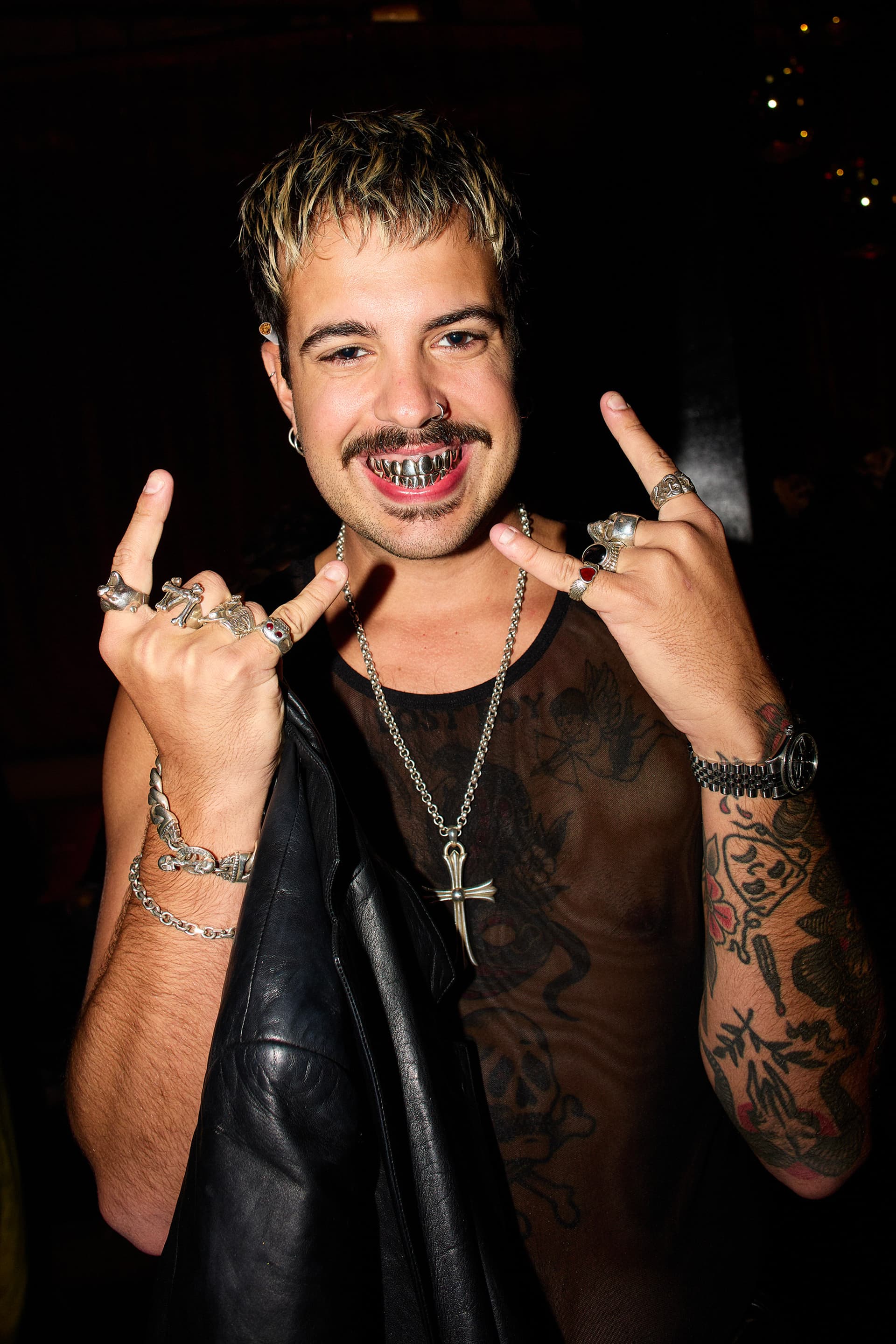 Man with facial piercings, silver grill teeth, tattoos, and rings showing rock hand signs wearing a sheer black top.