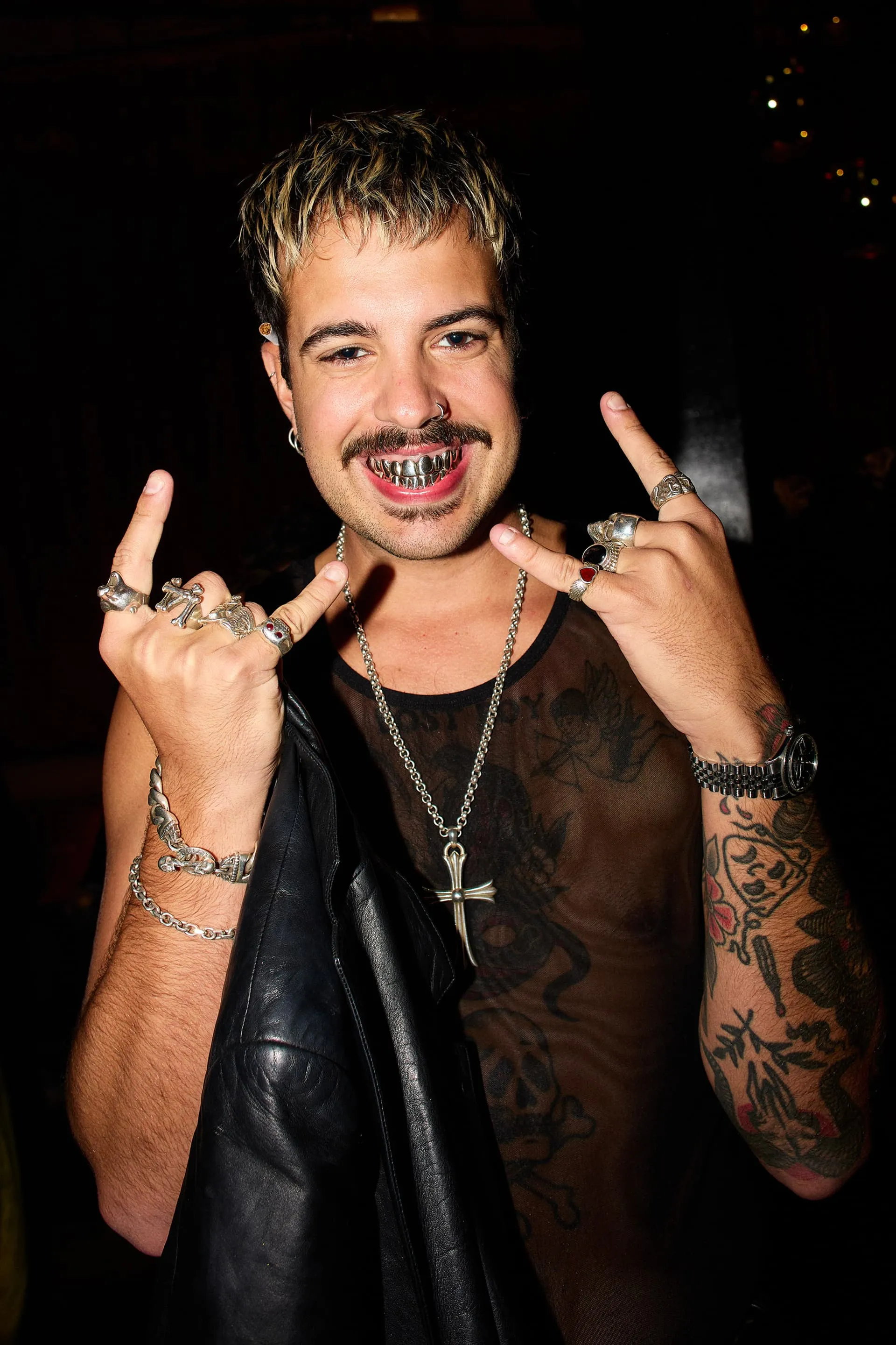 Man with facial piercings, silver grill teeth, tattoos, and rings showing rock hand signs wearing a sheer black top.