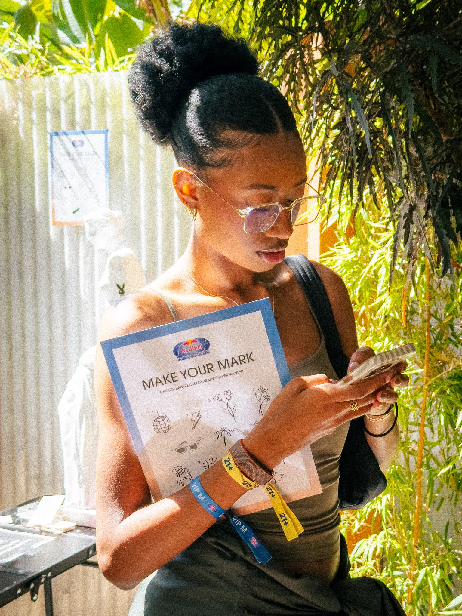 Young woman holding a 'Make Your Mark' flyer and looking at her phone outdoors with greenery.