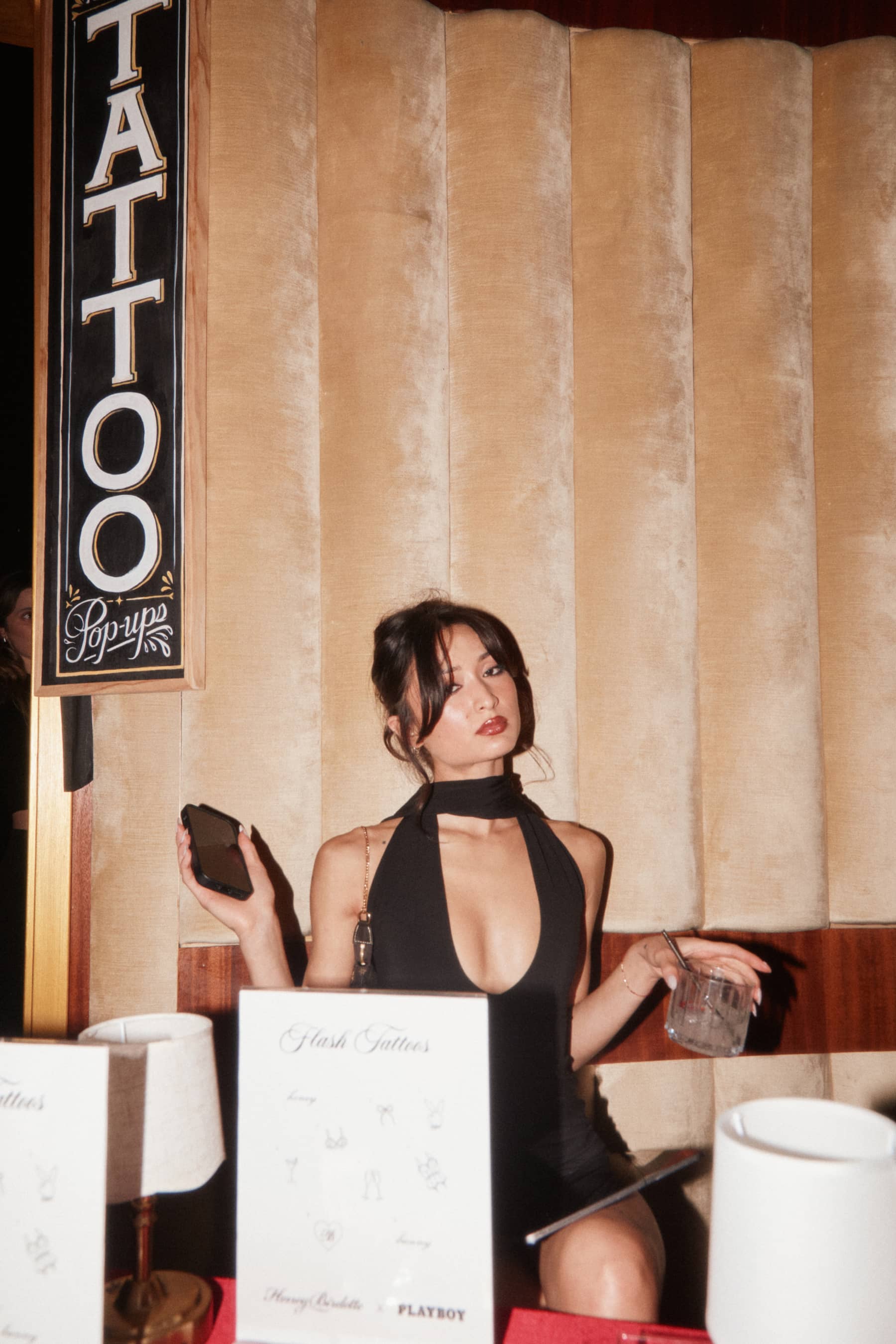 Woman in black dress sitting at a booth with a drink, phone, and tattoo flash menu at a pop-up.
