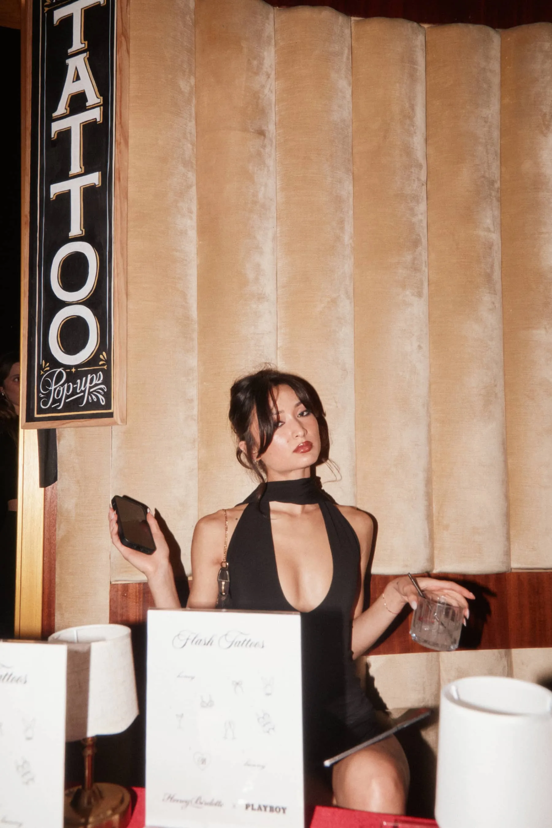 Woman in black dress sitting at a booth with a drink, phone, and tattoo flash menu at a pop-up.