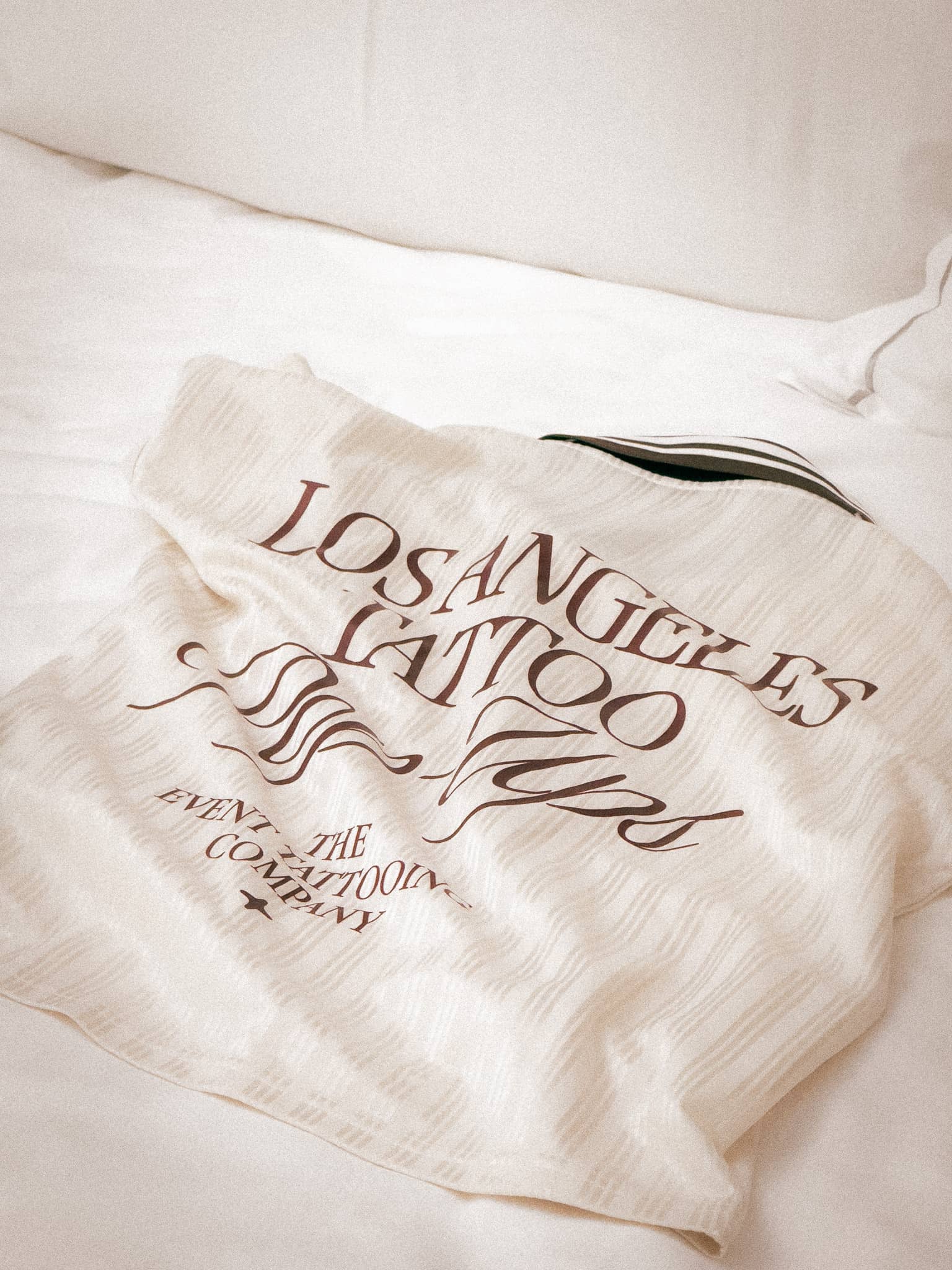 Beige jacket with text Los Angeles Tattoo Tours and The Tattooing Event Company laid on a white bed.