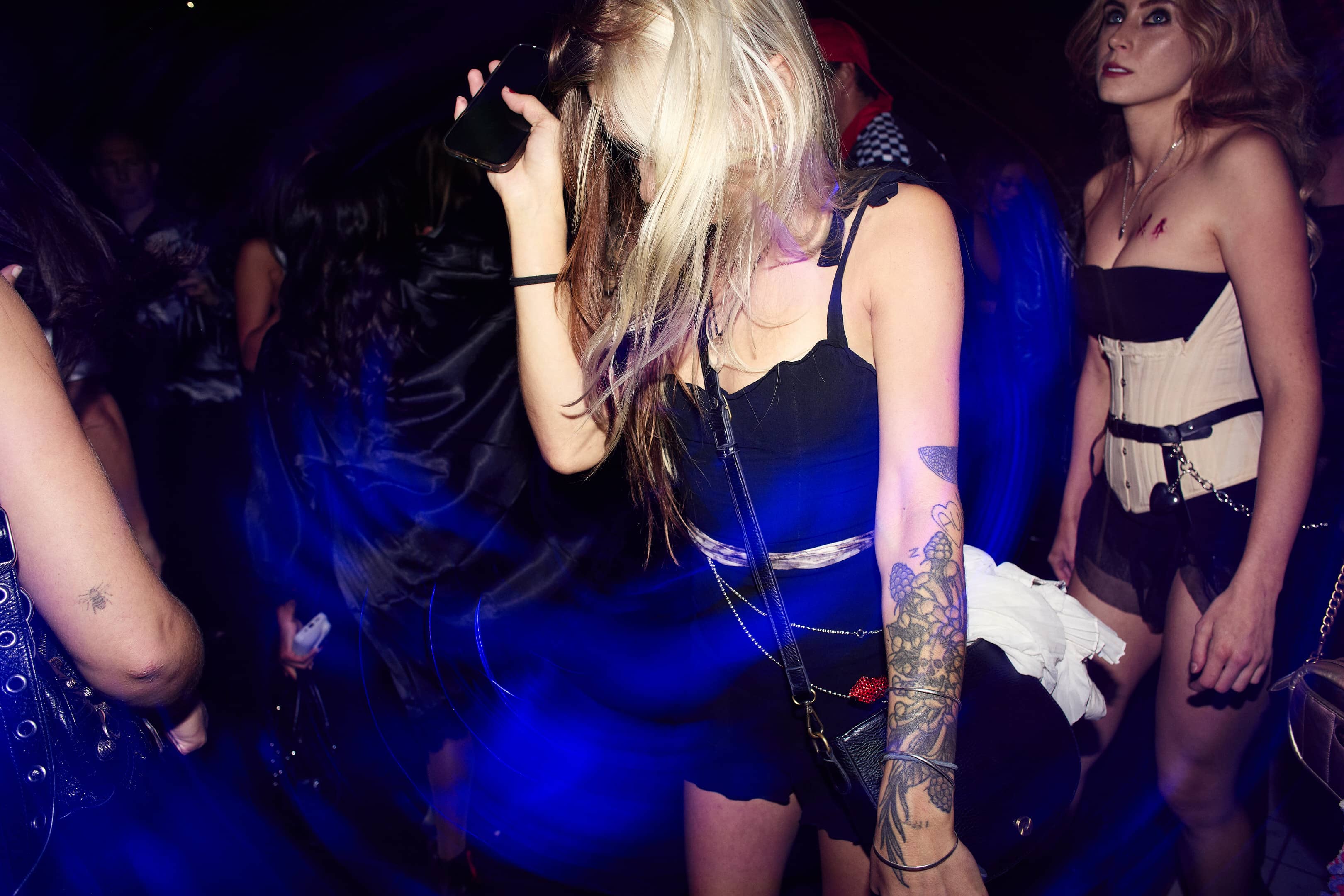 Young woman with floral tattoo dancing at a dim club with blue light trails, holding a phone near her face.