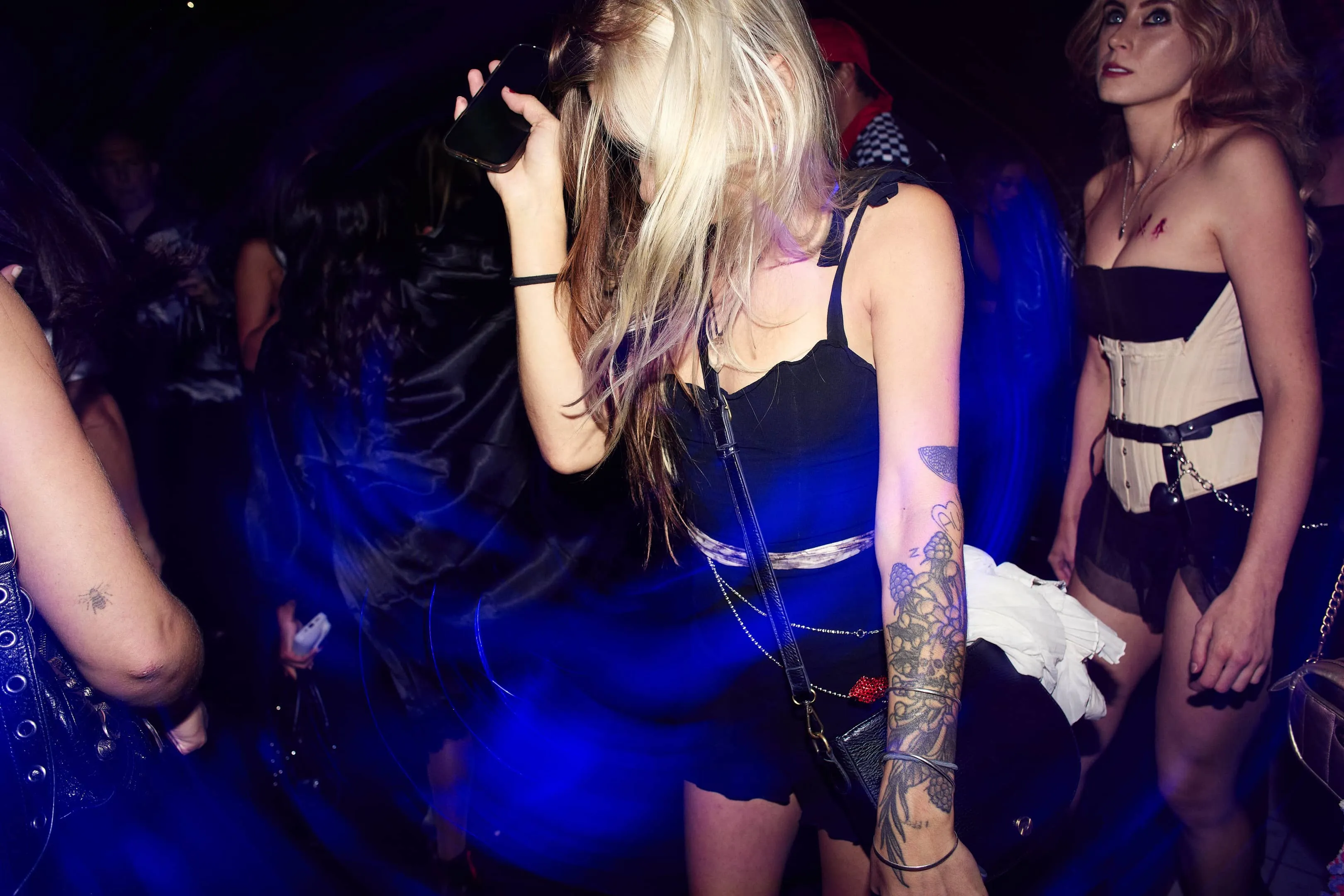 Young woman with floral tattoo dancing at a dim club with blue light trails, holding a phone near her face.
