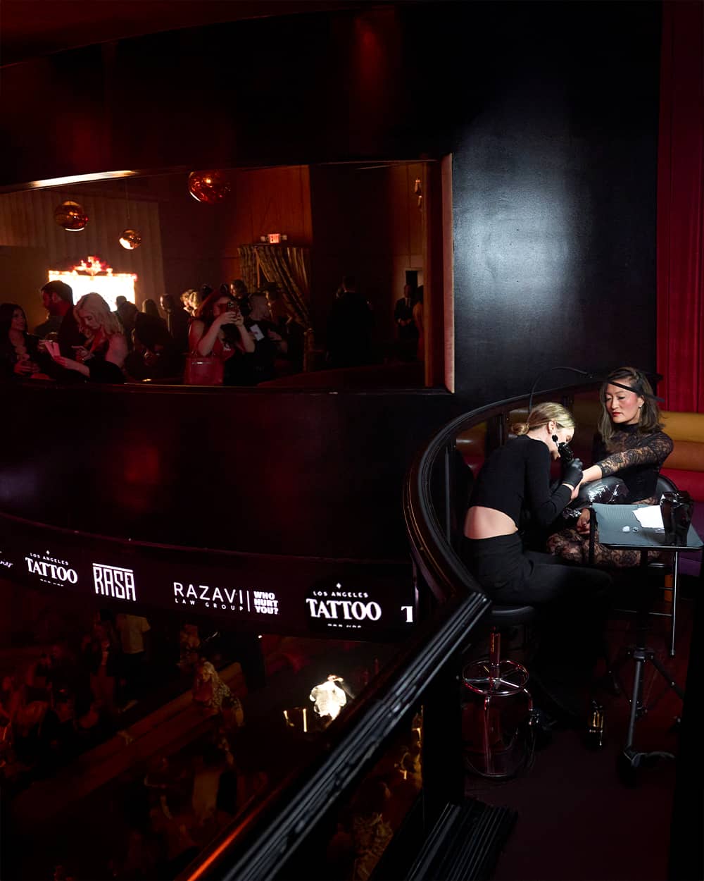 Tattoo artist wearing black works on a woman's arm in a dark, crowded nighttime venue.
