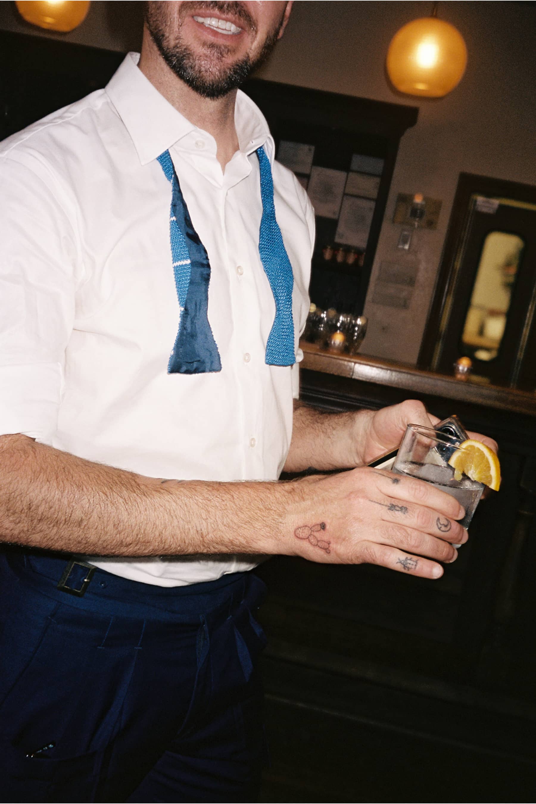 Man in white shirt with blue untied bow tie holding a drink with lemon in a dimly lit bar.