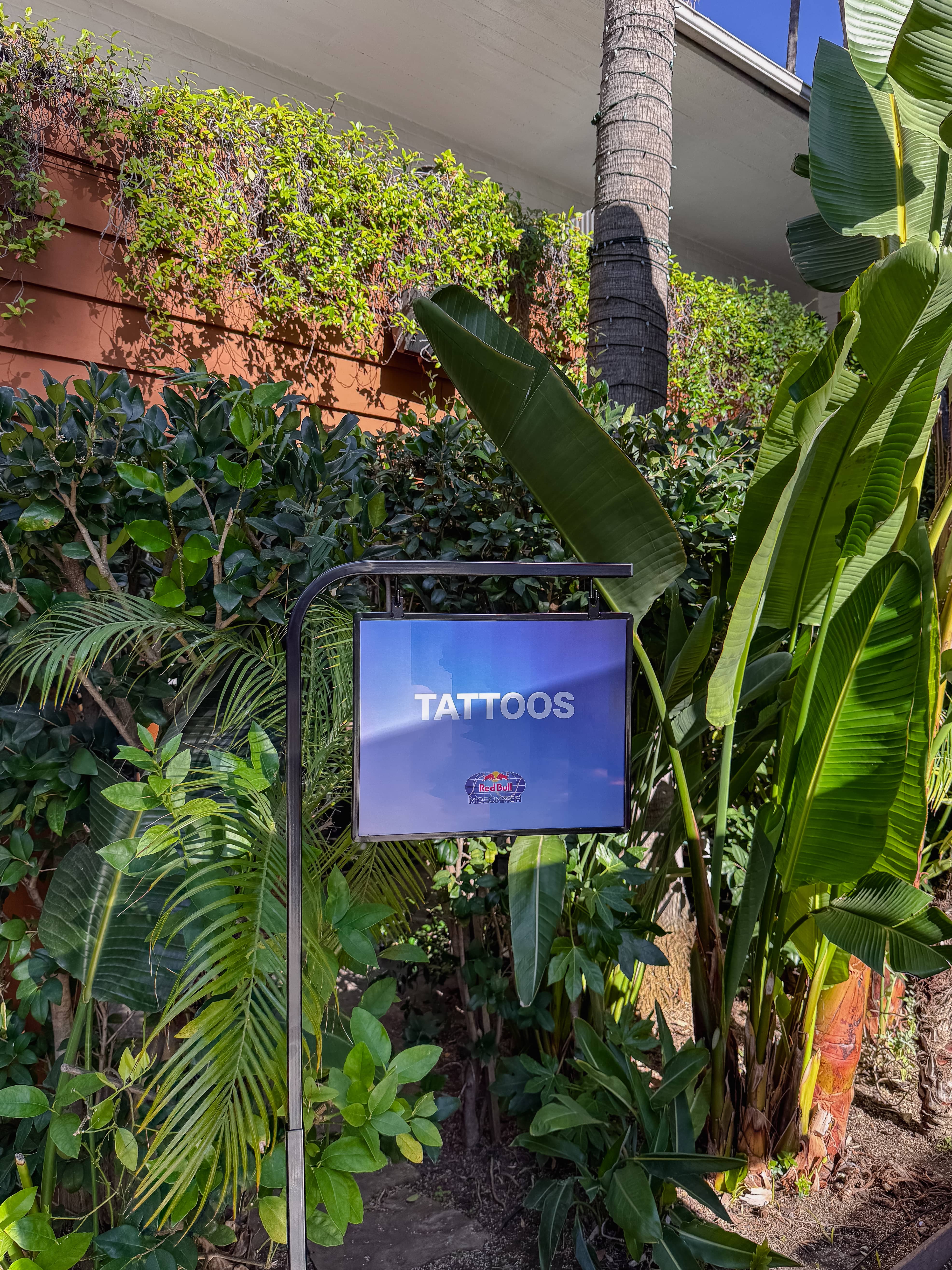 Blue sign with the word TATTOOS hanging on a metal stand among green plants outdoors.
