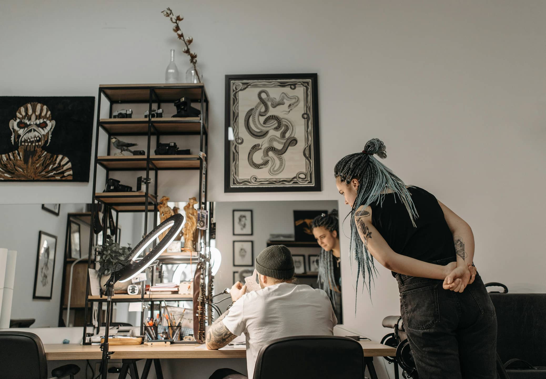 Tattoo artist with braided hair discussing a design with a seated client in a decorated studio.