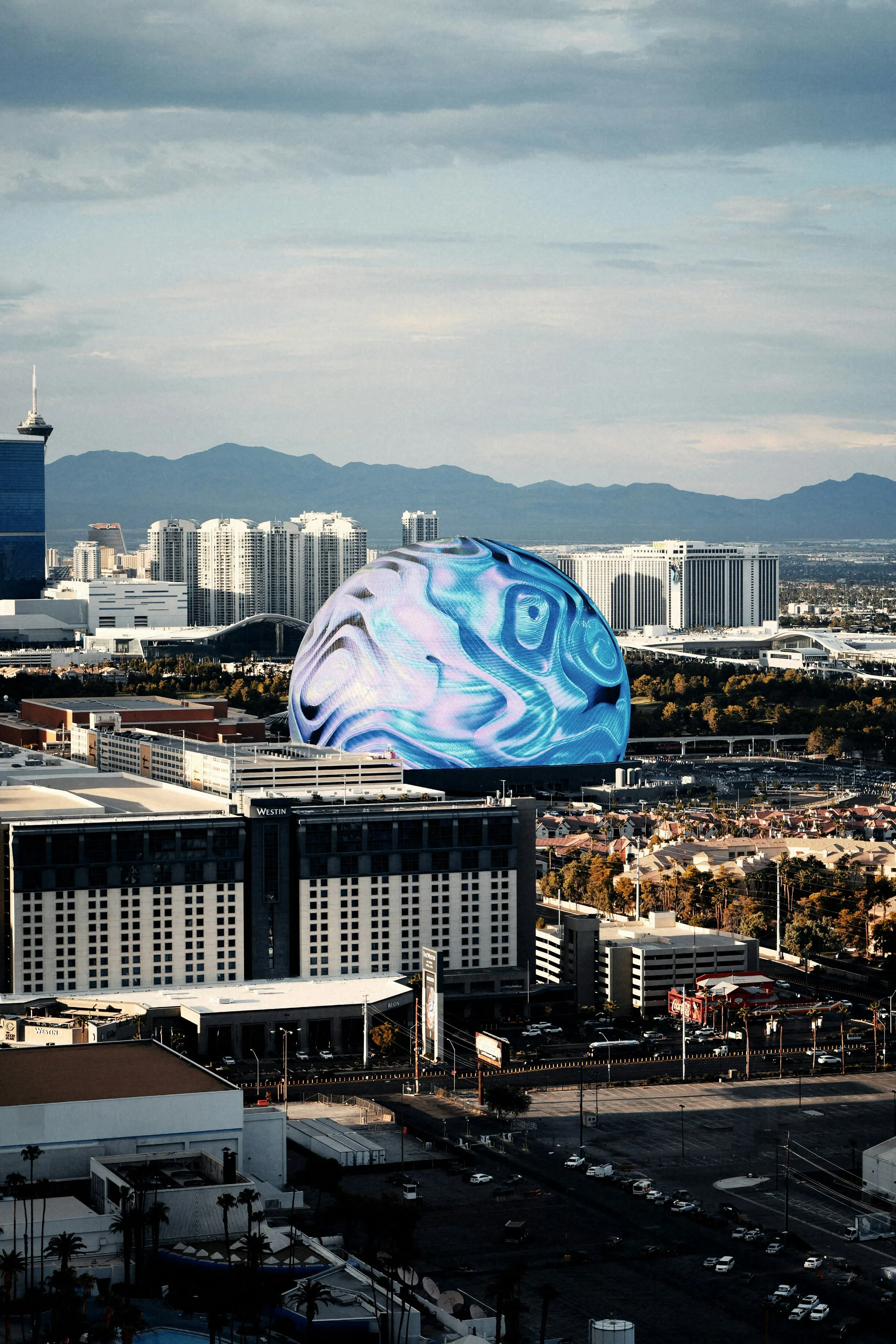 Large spherical display building with blue abstract design amid city buildings under a cloudy sky.