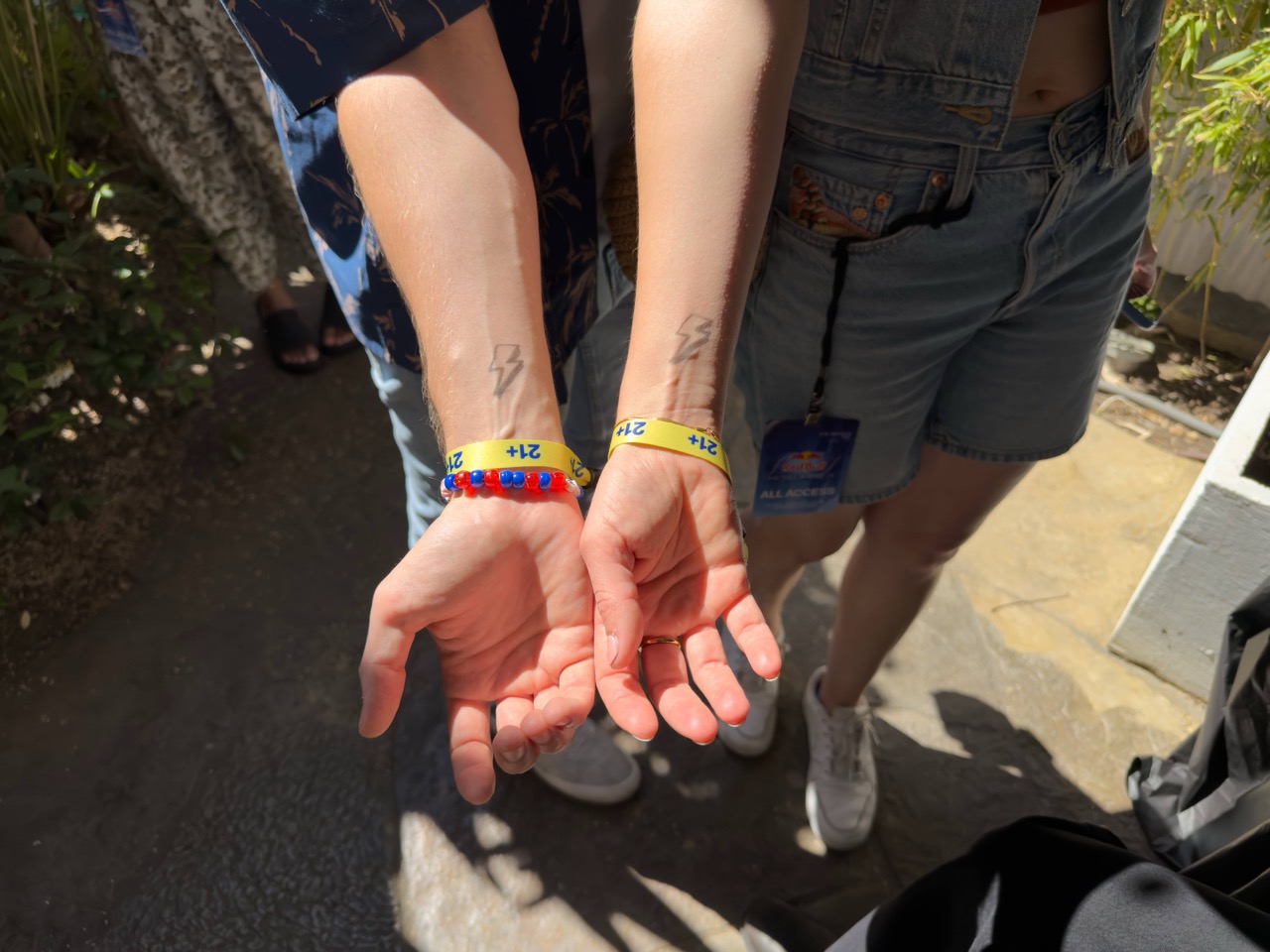Two forearms with matching lightning bolt tattoos and yellow 21+ wristbands in sunlight.