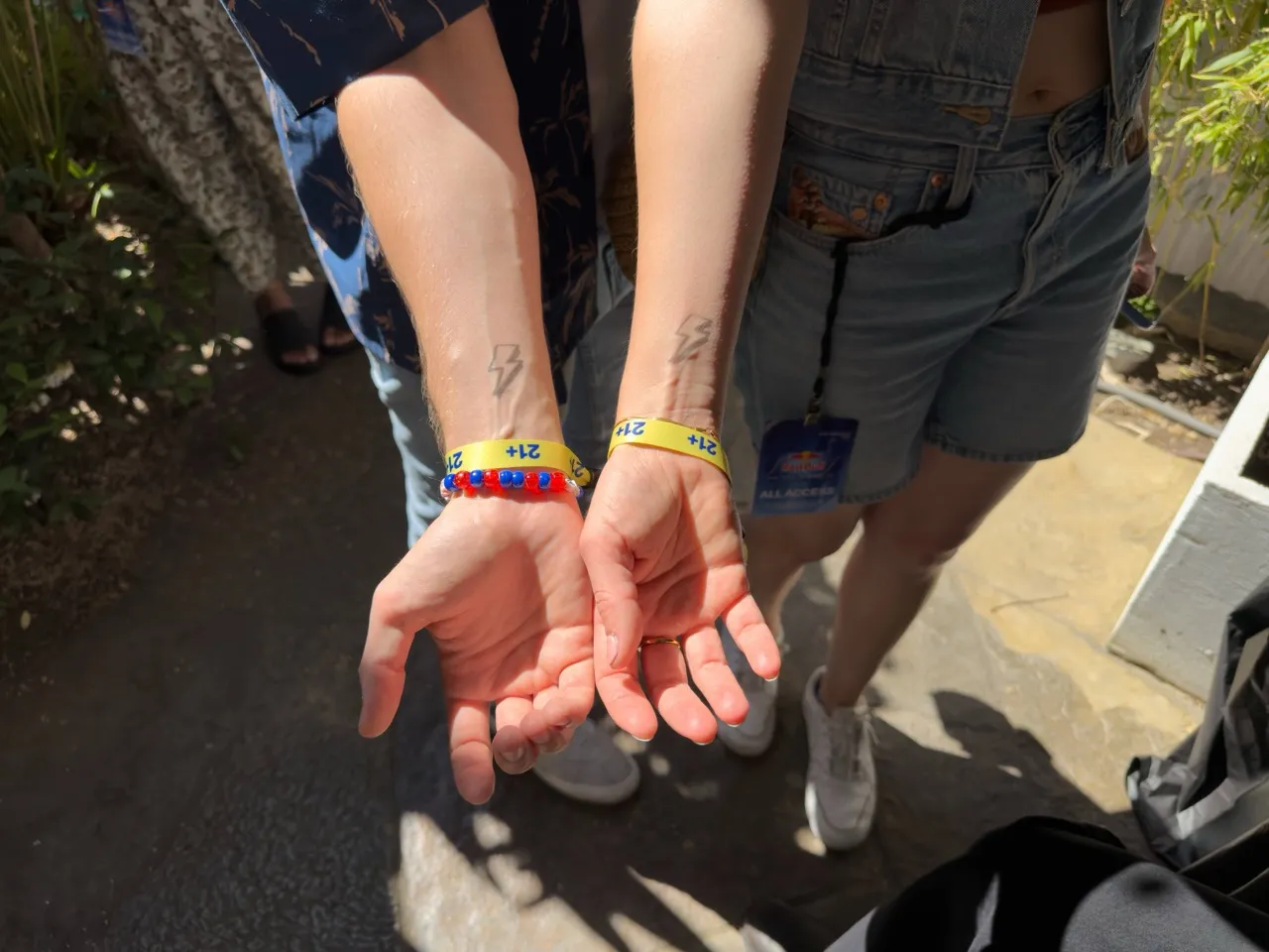 Two forearms with matching lightning bolt tattoos and yellow 21+ wristbands in sunlight.