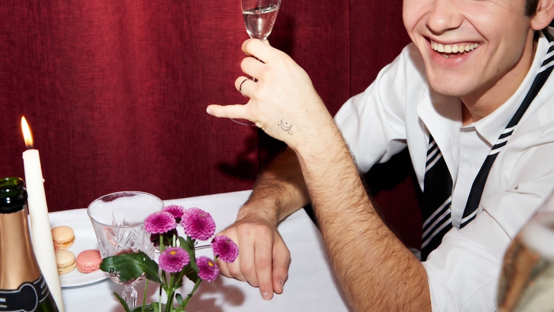 Smiling man in white shirt raises glass with moon tattoo on hand at a table with flowers and candle.