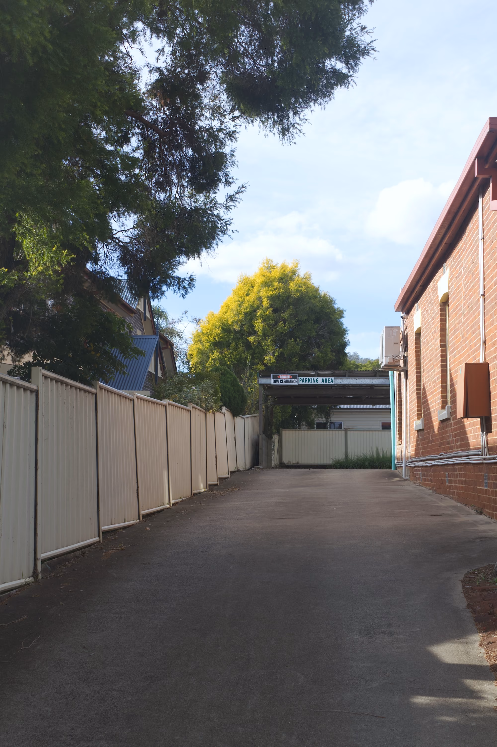 Driveway with a metal fence on the left, brick building on the right, and parking area sign ahead.