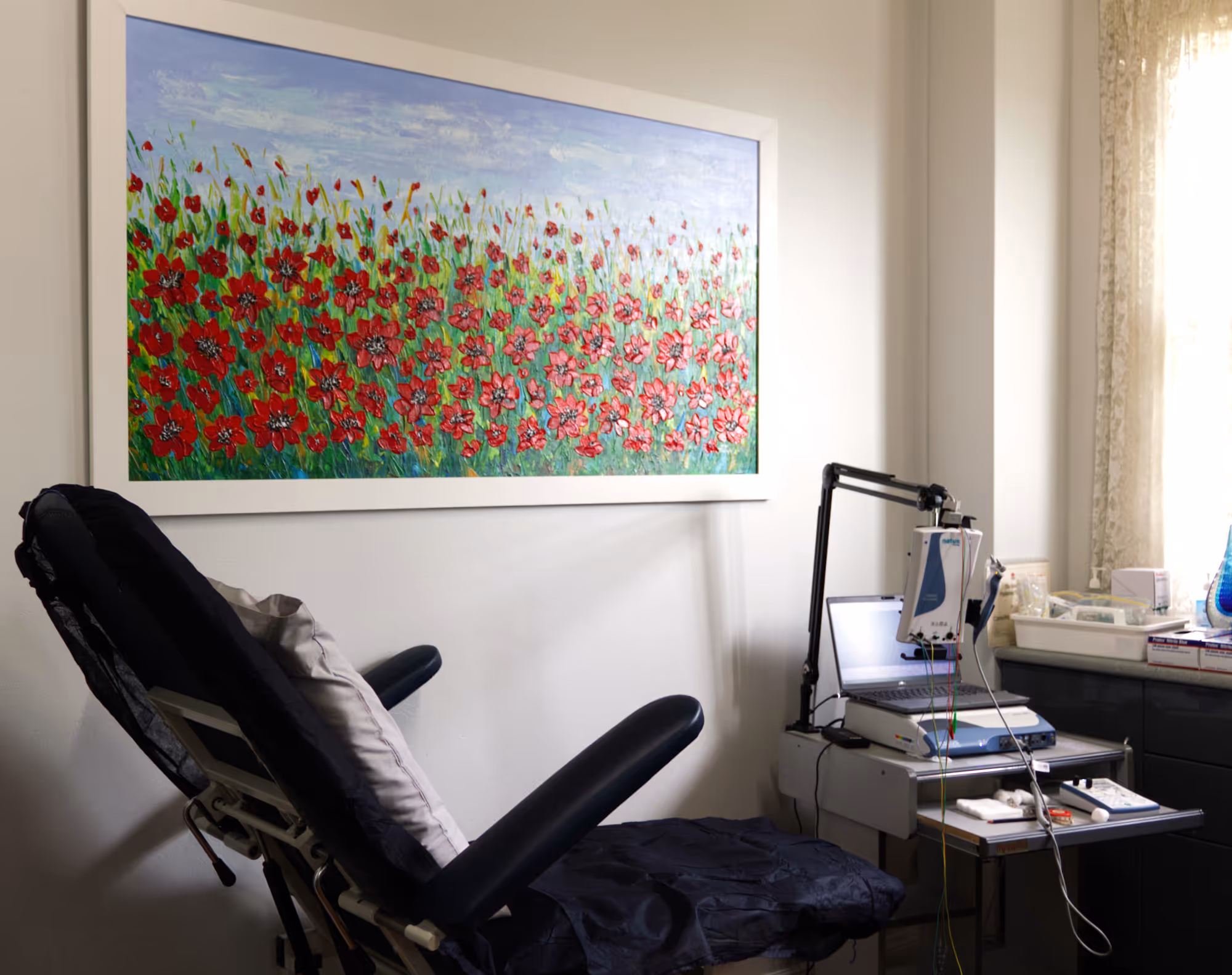 Neurology consultation room with medical chair, equipment, and bright floral painting on wall.