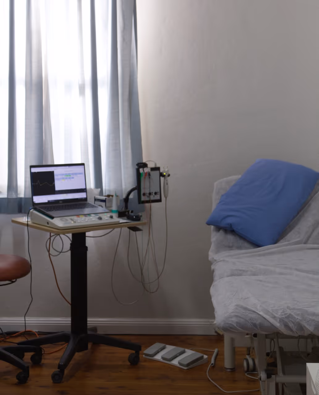 Medical examination room with a patient bed, blue pillow, equipment on a wheeled table, and monitor displaying graphs.
