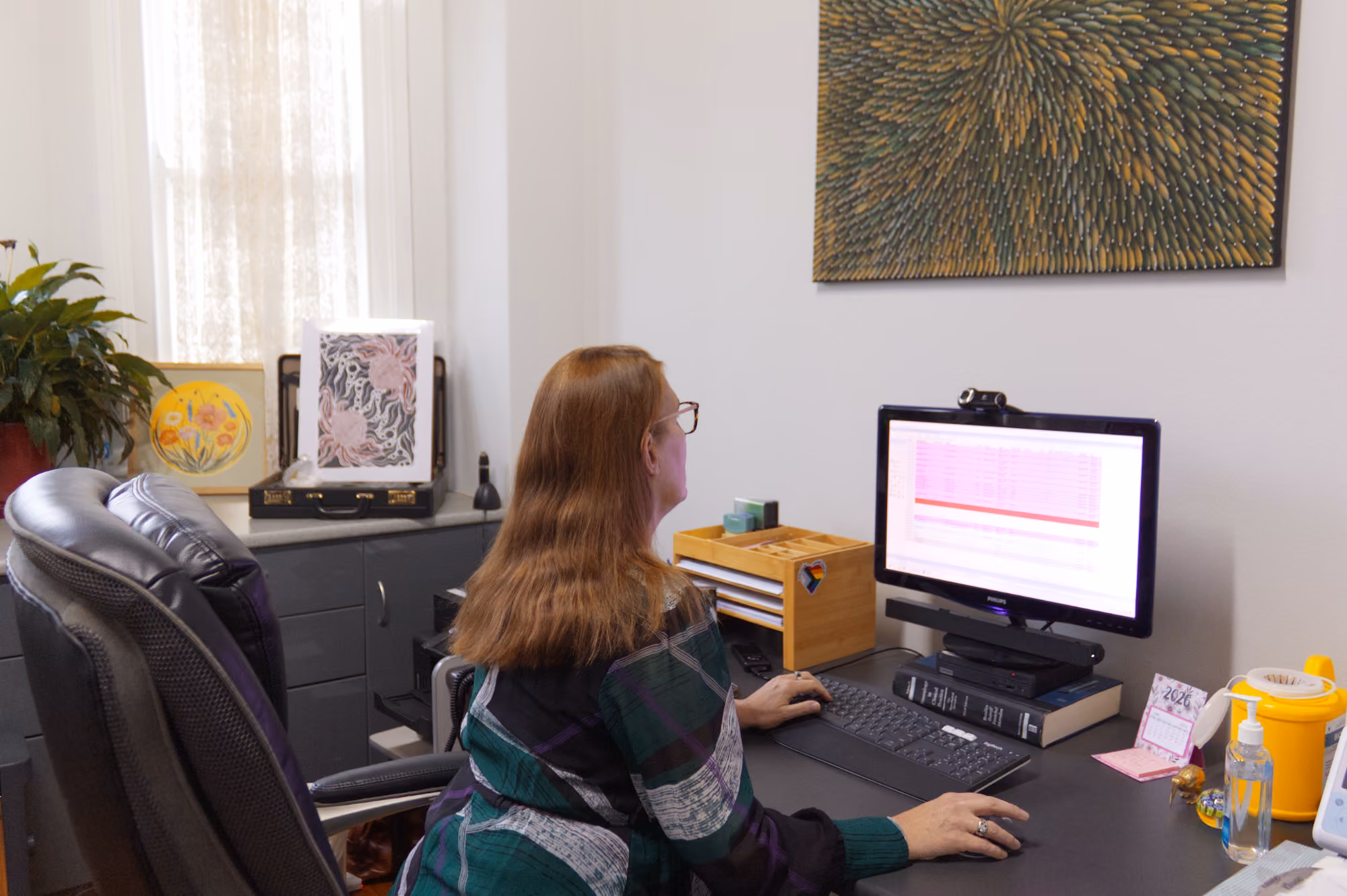 Woman with glasses working on a desktop computer in an office with plants and artwork.