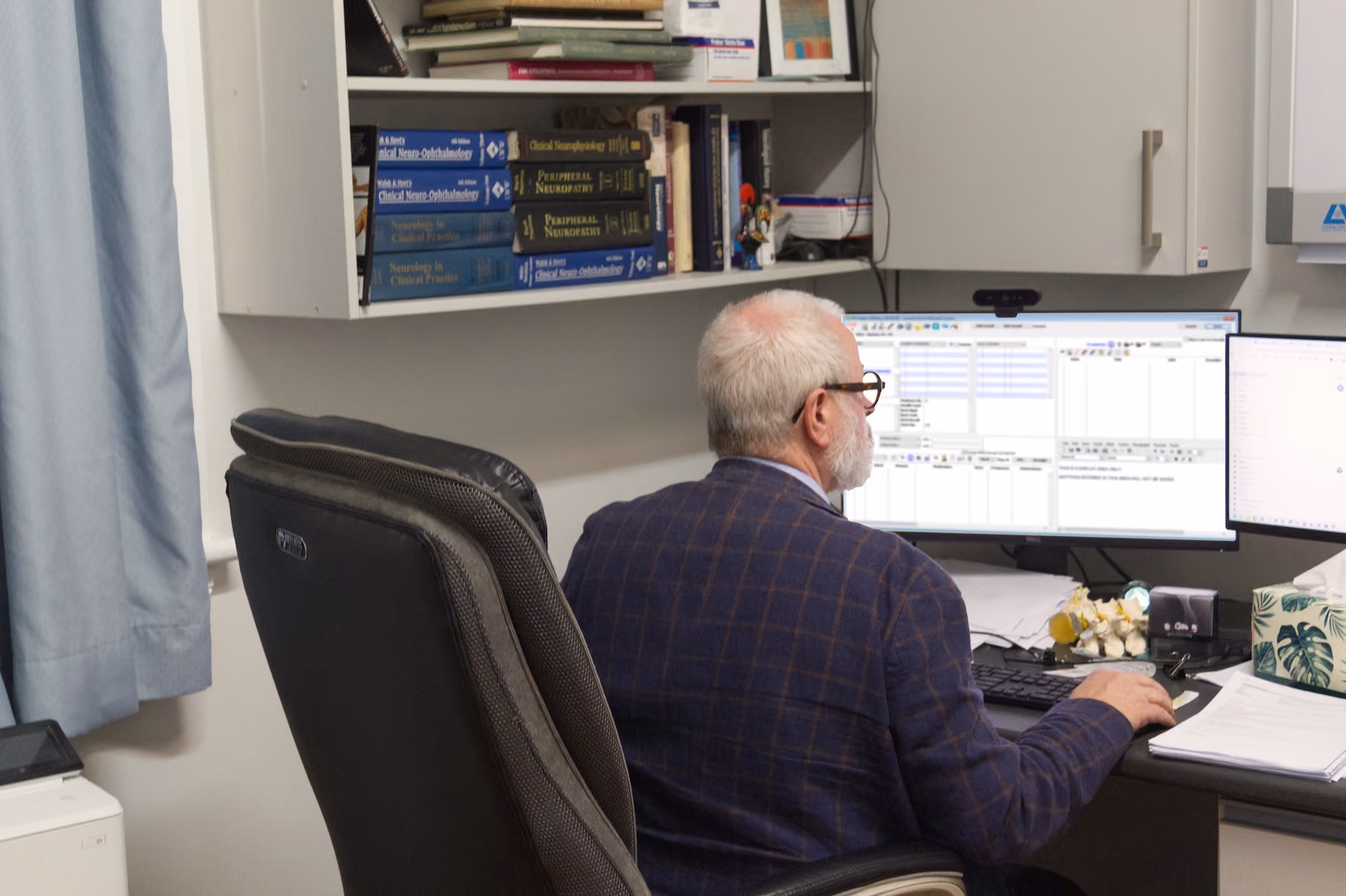 Older man with glasses working at a desk with dual monitors in an office with medical books on shelves.