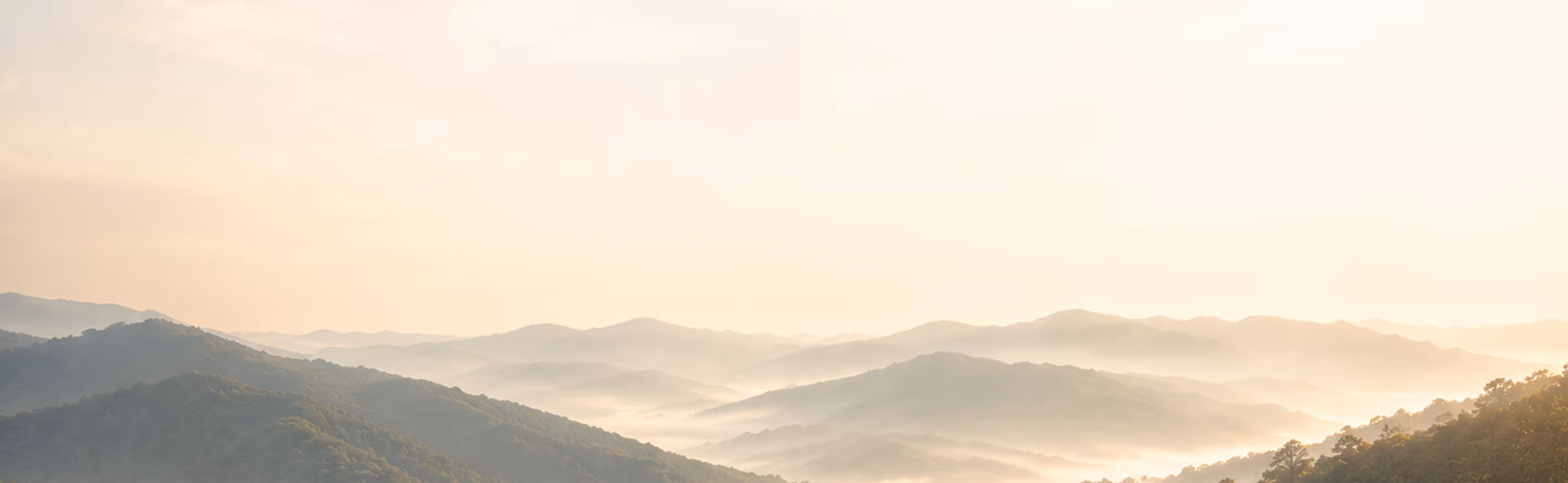 Layered mountain ranges fading into mist under a soft, bright sky at sunrise or sunset.