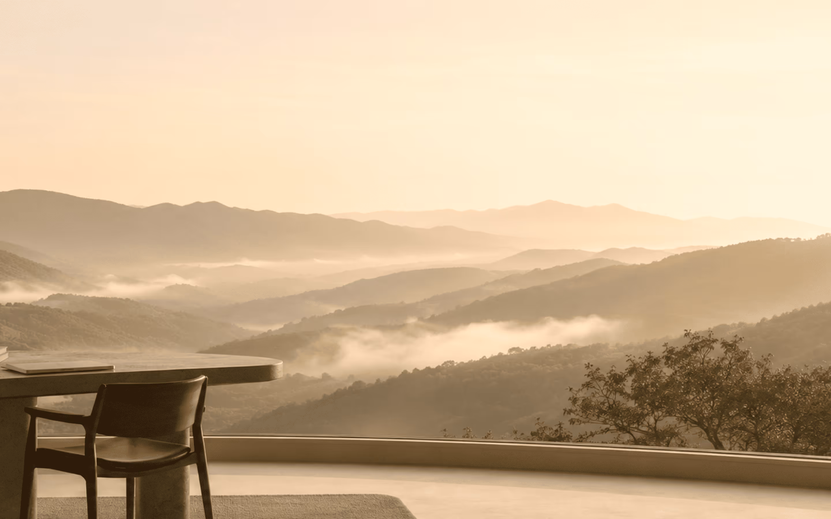 Wooden chair and table inside with a window view of misty layered mountain hills at sunrise.