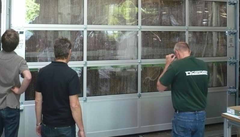 Drei Männer stehen vor einem großen Industrie-Sektionaltor und betrachten es aufmerksam. Das Tor besteht aus einem hellen Metallrahmen (vermutlich Aluminium) und ist größtenteils mit großflächigen, transparenten Elementen ausgestattet, durch die sich die Umgebung spiegelt. Der Mann auf der rechten Seite trägt ein dunkelgrünes T-Shirt mit dem weißen Schriftzug „ConDoor“ auf dem Rücken.