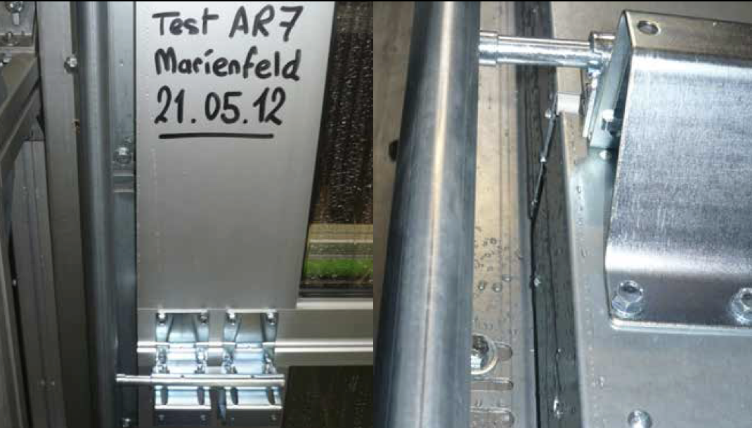 Zweigeteiltes Bild, das Details eines industriellen Tests zeigt. Auf der linken Seite ist ein Aluminiumrahmen abgebildet, auf dem handschriftlich mit schwarzem Stift "Test AR7", "Marienfeld" und das Datum "21.05.12" steht. Unten am Rahmen sind metallische Beschläge montiert. Die rechte Seite zeigt eine Nahaufnahme einer massiven Metallhalterung und einer Führungsschiene oder eines Rohrs. Auf den Metalloberflächen rechts sind deutlich Wassertropfen zu erkennen, was auf einen Dichtigkeits- oder Wassertest hinweist.