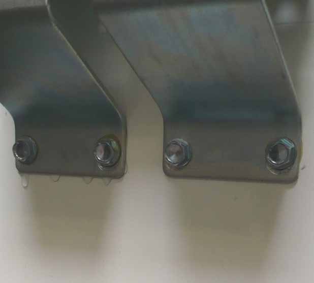Nahaufnahme von zwei metallischen Winkeln oder Beschlägen, die mit Sechskantschrauben an einer weißen, glatten Oberfläche befestigt sind. An der unteren Kante des linken Beschlags hängen deutlich sichtbare Wassertropfen herab, was auf Feuchtigkeit oder eine Leckage während eines Wasserdichtigkeitstests hinweist.