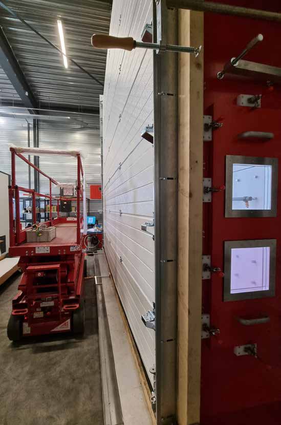 Seitenansicht eines großen, weißen Industrie-Sektionaltores, das für Tests an einer massiven roten Prüfkammer montiert ist. Das Tor ist in einer Führungsschiene fixiert und mit Schraubzwingen an einem Holzrahmen befestigt. An der roten Prüfwand sind kleine quadratische Sichtfenster zu erkennen. Im Hintergrund der Halle steht eine eingefahrene rote Scherenarbeitsbühne.