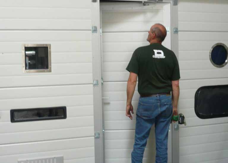 Rückansicht eines Mannes in Jeans und einem dunkelgrünen T-Shirt mit dem weißen Logo „ConDoor“. Er steht vor einem weißen, gerippten Industrie-Sektionaltor und blickt nach oben, um die obere Kante einer integrierten, leicht geöffneten Schlupftür (Gehtür) zu inspizieren. Das Sektionaltor ist links und rechts neben der Tür mit verschiedenen Fenstertypen (eckig, länglich, rund) versehen.
