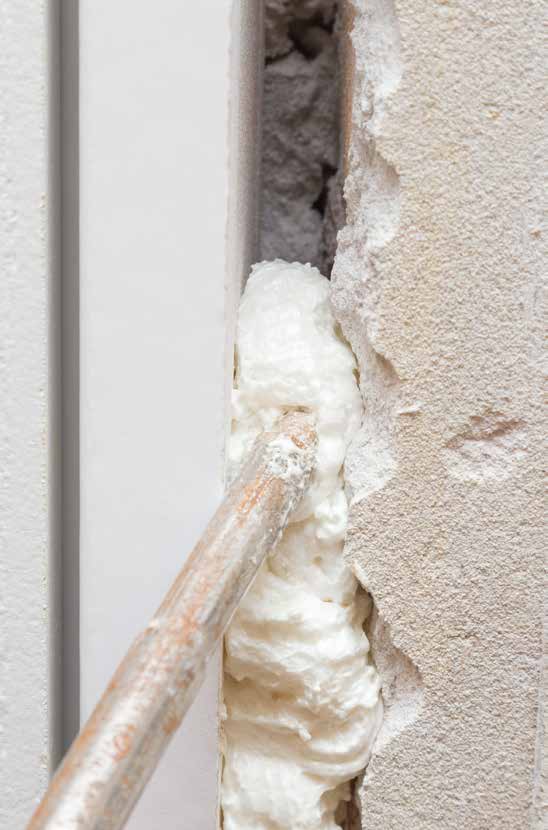 Nahaufnahme der Ausfüllung einer schmalen Baufuge zwischen einem weißen Profil und einer rau verputzten Wand. Ein langes Metallspritzrohr trägt frischen, hellen PU-Bauschaum (Montageschaum) in den Hohlraum ein, um die Fuge abzudichten und zu dämmen.