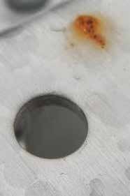 Nahaufnahme einer hellgrauen, leicht strukturierten metallischen Oberfläche. Im unteren linken Bereich ist ein sauberes, kreisrundes Loch (eine Bohrung oder Stanzung) zu sehen. Etwas weiter rechts oberhalb davon befindet sich ein deutlich sichtbarer, orange-brauner Rostfleck, der auf eine beginnende lokale Korrosion des Materials hindeutet.