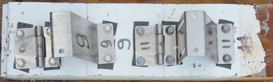 Aufsicht auf eine weiße Materialprobe, vermutlich ein Abschnitt eines Sektionaltorpaneels, auf der zwei Gruppen von metallischen Beschlägen festgeschraubt sind. Jede Gruppe besteht aus einem flachen Scharnier und einem abgewinkelten Beschlagteil. Die Metallteile und der Untergrund sind handschriftlich mit schwarzem Stift markiert: Die linke Baugruppe trägt mehrfach die Nummer „9“, die rechte die Nummer „11“. Unter den Verschraubungen liegen schwarze Unterlegpads, und an den äußeren Rändern der weißen Platte sind bräunliche Verfärbungen zu erkennen.