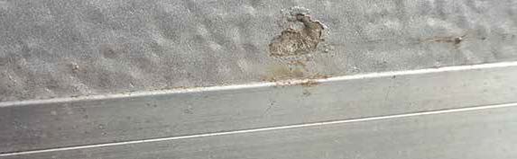 Nahaufnahme der grauen, geprägten Oberfläche (sogenannte Stucco-Struktur) eines metallischen Torpaneels. Im oberen Bereich der Struktur befindet sich ein deutlicher Lackabplatzer, an dem die äußere Beschichtung fehlt und das darunterliegende, leicht bräunlich angerostete Material freiliegt. Direkt unterhalb dieser Beschädigung schließt sich ein glattes, horizontales Metallprofil an.