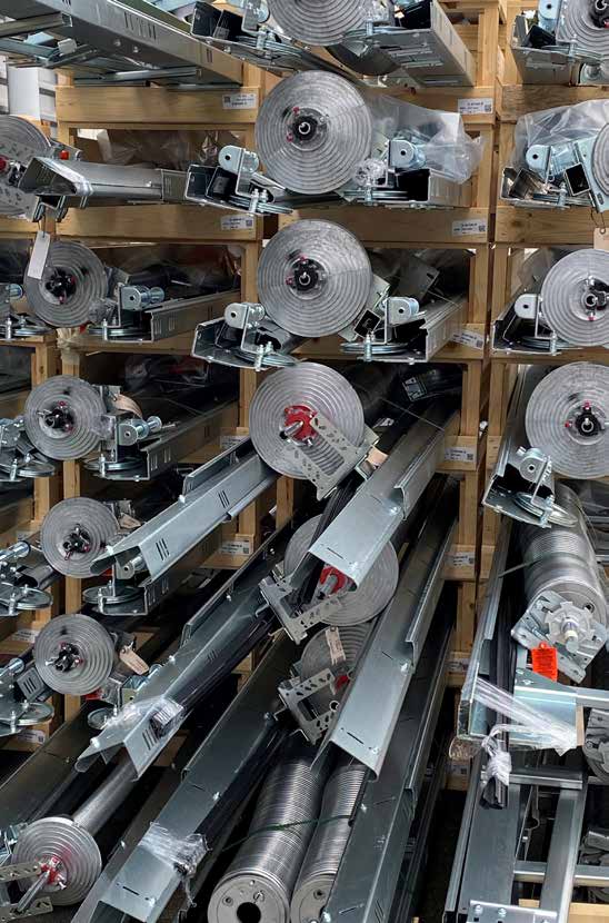 Blick auf ein dicht bepacktes Regalsystem aus Holz, in dem zahlreiche metallische Bauteile für Industrietore gelagert werden. Eng übereinander und nebeneinander gestapelt liegen dort vormontierte Baugruppen aus verzinktem Stahl. Besonders auffällig sind die massiven, spiralförmigen Torsionsfedern mit integrierten Seiltrommeln und Halterungen sowie die langen, U-förmigen Führungsschienen. Einige Endstücke der Federmechanik weisen rote und schwarze Markierungen auf.