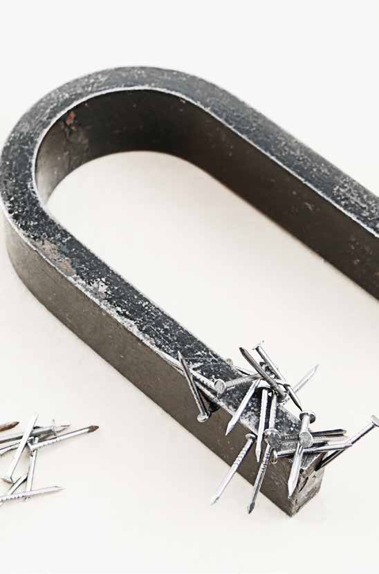 Nahaufnahme eines großen, massiven Hufeisenmagneten aus dunklem, leicht abgenutztem Metall, der auf einem hellen Untergrund liegt. An seinem rechten Pol haften viele kleine, silberne Stahlnägel kreuz und quer aneinander, die durch die Magnetkraft angezogen werden. In der unteren linken Bildhälfte liegt ein weiteres, kleines Häufchen solcher Nägel lose auf dem Untergrund.