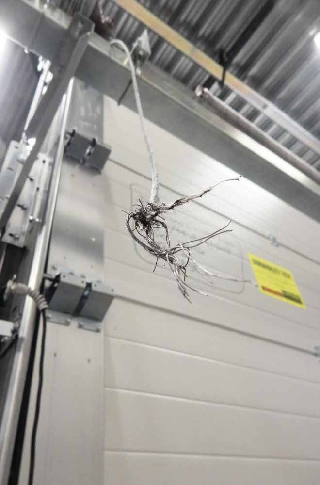 Nahaufnahme eines gerissenen Stahlseils, das von der Deckenkonstruktion eines Sektionaltores herabhängt. An der Bruchstelle ist das Seil extrem stark ausgefranst; die unzähligen dünnen Drahtlitzen stehen wirr und aufgesplisst in alle Richtungen ab. Im leicht unscharfen Hintergrund sind die weißen, horizontalen Paneele eines geschlossenen Industrietores sowie die massiven metallischen Führungsschienen auf der linken Seite zu erkennen, was verdeutlicht, dass es sich um das gerissene Tragseil der Toranlage handelt.