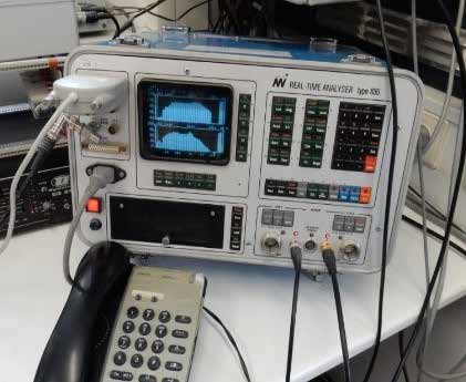 Fotografie eines elektronischen Messgeräts, eines „Real-Time Analyser type 830“ in einem hellgrauen Gehäuse, das auf einem Tisch steht. Auf dem eingebauten Bildschirm sind blaue grafische Diagramme oder Frequenzspektren zu erkennen. Das Gerät ist mit zahlreichen Tastenfeldern, Schaltern und angeschlossenen Messkabeln ausgestattet. Im Vordergrund davor befinden sich das Tastenfeld und der Hörer eines Tischtelefons.