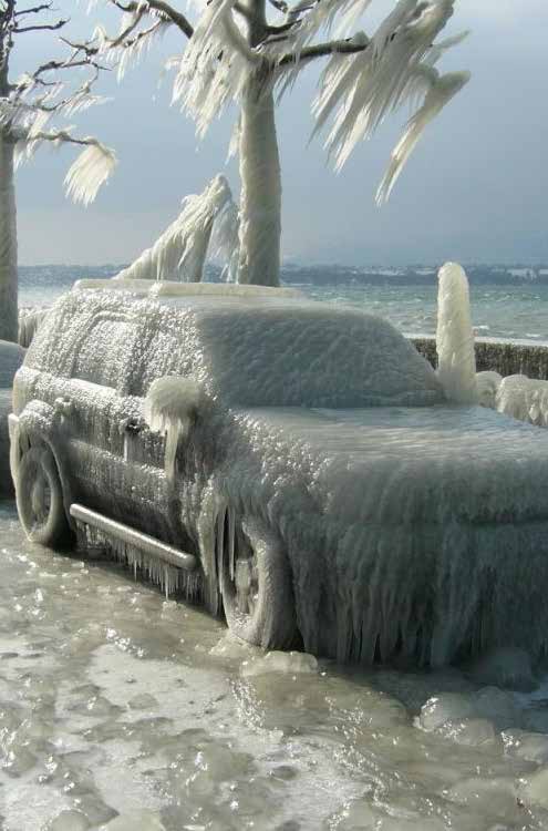 Fotografie eines Autos, das komplett von einem dicken Eispanzer und langen Eiszapfen umschlossen ist. Im Hintergrund sind ebenfalls stark vereiste Bäume und ein Uferbereich an einem Gewässer zu sehen, was auf extreme Winterbedingungen mit gefrierender Gischt hindeutet.