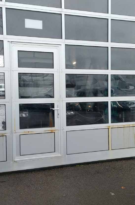 Außenansicht eines Sektionaltores mit integrierter Schlupftür (Personentür), einer Aluminium-Rahmenkonstruktion sowie großen Glas- und Paneelelementen, an dessen unteren Metallteilen leichte Rostspuren zu erkennen sind.