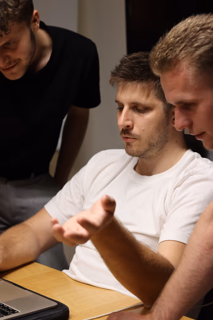 Three men collaborate and discuss while looking at a laptop on a wooden table.