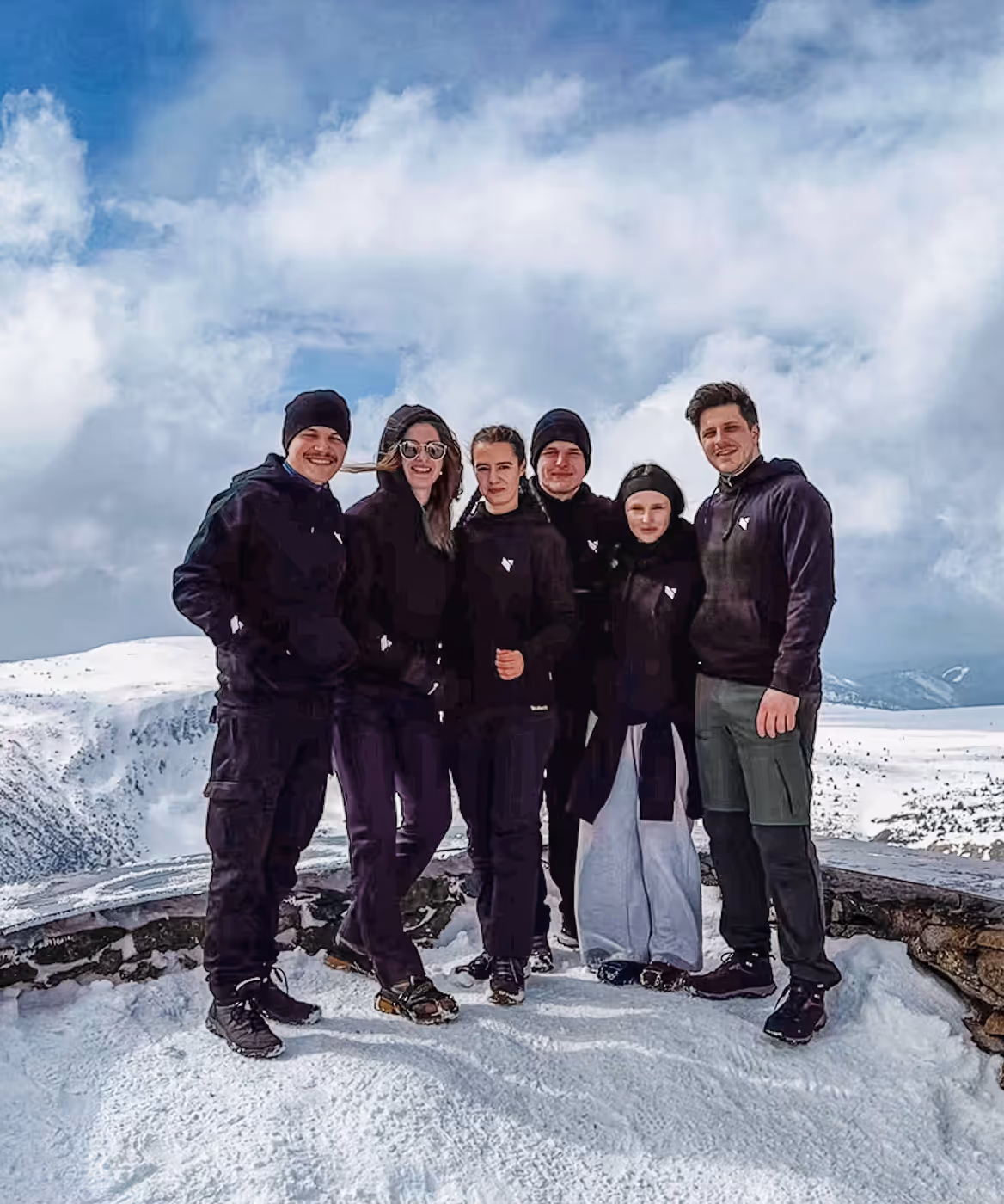 Group of six people standing together on snow with mountains in the background under a cloudy sky.
