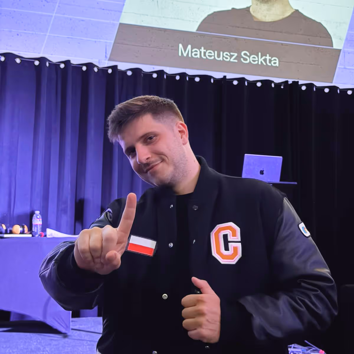 Young man with short hair in a black jacket showing index finger, with black curtain and screen behind.