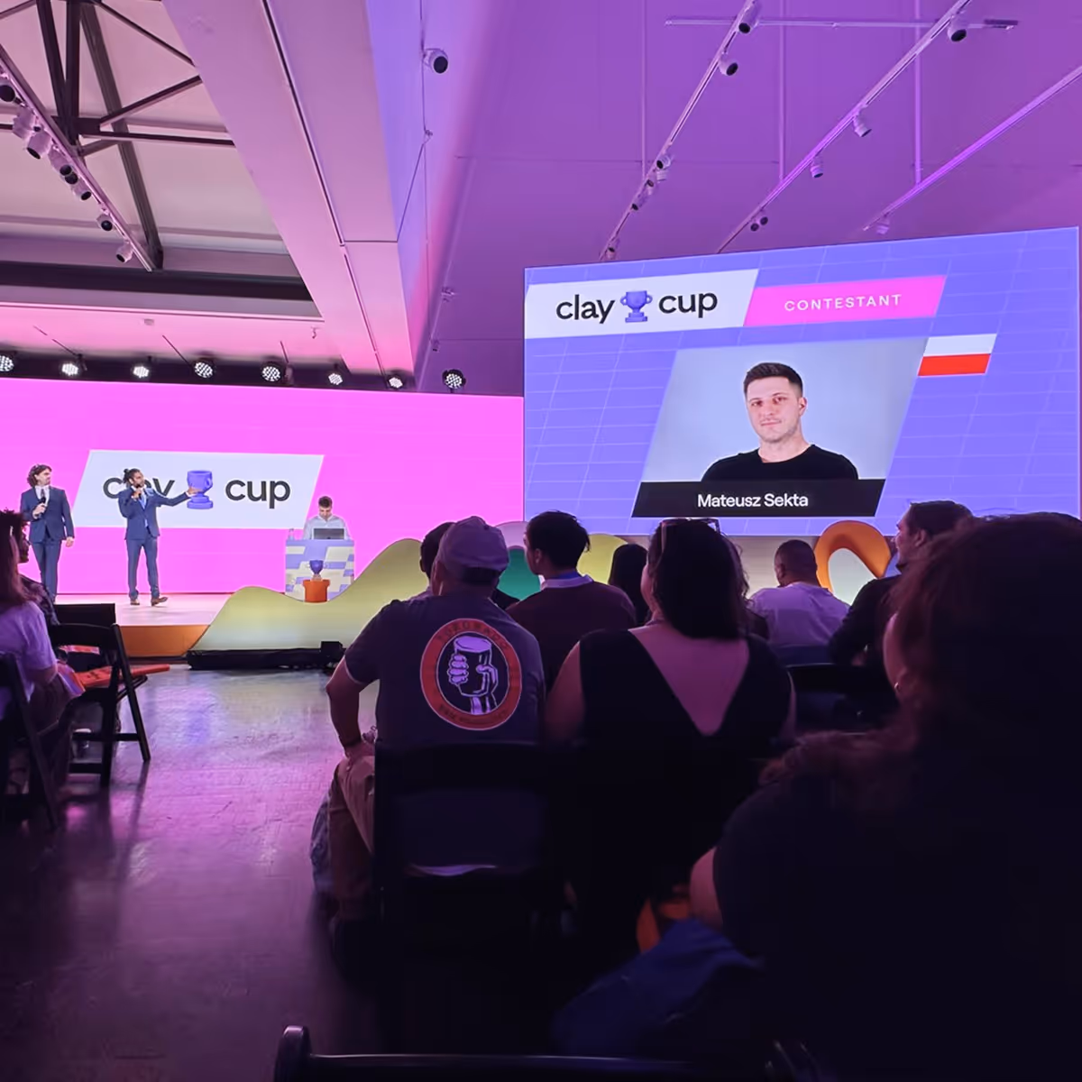Audience watching Clay Cup contest with two hosts on stage and contestant Mateusz Sekta on screen.